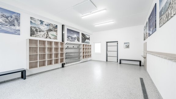 A bright, modern room with gray flooring and white walls. Large, impressive pictures of mountains hang on the walls, and the room is furnished with shelves and benches. - Appartementhaus Kathrin - Sölden