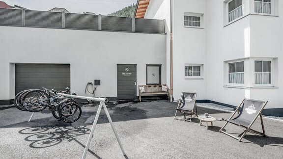 A modern courtyard with bicycle racks and lounge chairs. The walls are white, and there is a garage door in the background. - Appartementhaus Kathrin - Sölden