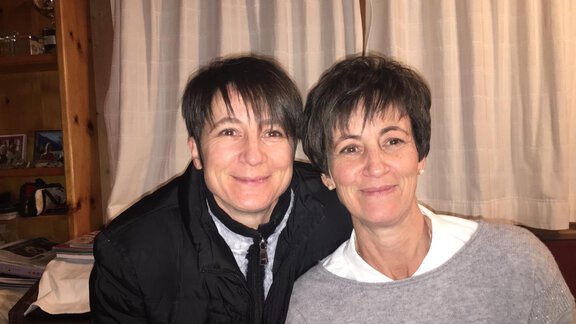 Two women smile kindly in front of a background with curtains and wooden shelves. Both are wearing simple, comfortable clothing and appear relaxed. - Gästehaus Martin - Sölden