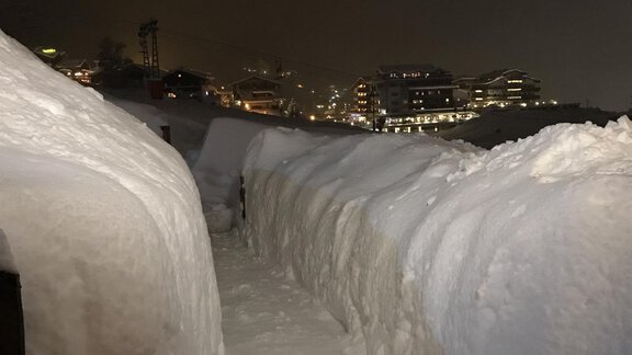 A snowy path between high snow walls at night. In the background, lights of buildings are visible. - Gästehaus Martin - Sölden