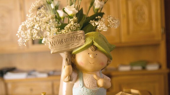 A small figure of a boy with a sign saying "Welcome." In the background, fresh flowers are in a vase. - Hotel Sportpension Carinthia - Sölden