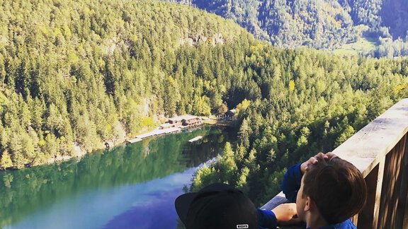 Two boys are sitting at a viewpoint, looking at a clear river surrounded by green mountains. The nature is lush and enchanting. - Hotel Sportpension Carinthia - Sölden