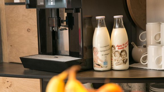 A modern coffee machine is on a wooden shelf. Next to the machine, there are two bottles of milk, and in the foreground, a selection of fresh fruit is visible. - Hotel Sportpension Carinthia - Sölden