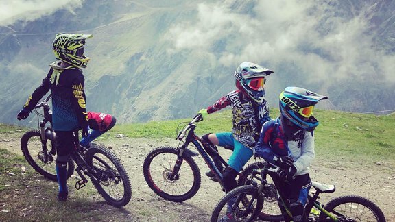Three children are standing with their mountain bikes on a mountain. In the background, clouds and impressive mountains can be seen. - Hotel Sportpension Carinthia - Sölden