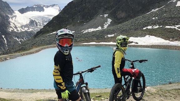 Two young cyclists are standing next to a clear blue lake in the mountains. In the background, snow-capped peaks and a beautiful landscape can be seen. - Hotel Sportpension Carinthia - Sölden
