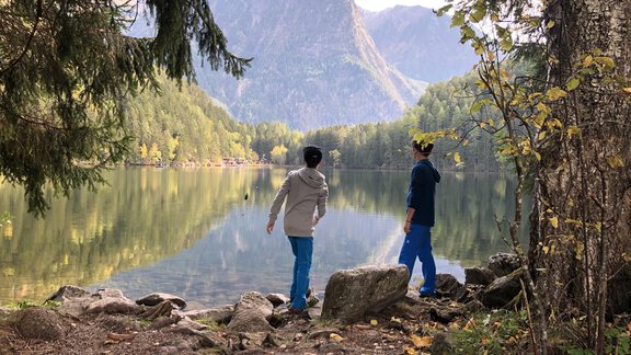 Two people are standing at the shore of a calm lake, looking at the mountains in the background. The surroundings are characterized by trees and autumn colors. - Hotel Sportpension Carinthia - Sölden