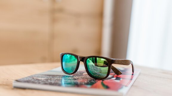 A pair of sunglasses with green lenses rests on a magazine. The background is bright and neutral. - Hotel Sportpension Carinthia - Sölden