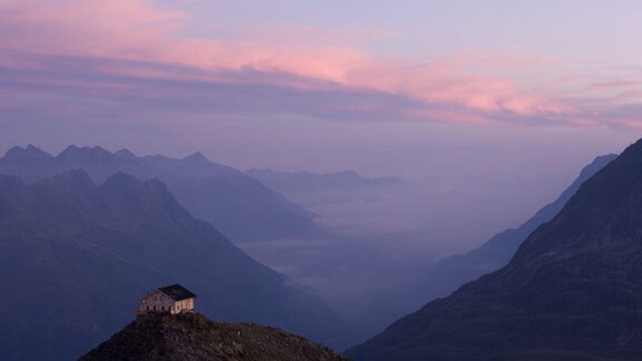 A small cabin stands on a hill in the mountains. The sky is tinted in soft shades of purple and pink. - Landhaus Falkner - Sölden