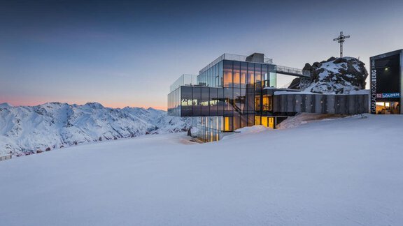 A modern building made of glass and concrete on snow-covered terrain. Majestic mountains rise in the background under a clear sky. - Landhaus Falkner - Sölden