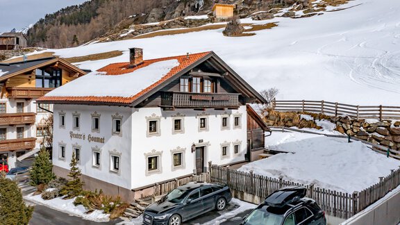 A traditional white building with a red tile roof stands in front of a snow-covered landscape. Cars are parked in front of the house, surrounded by a tranquil environment. - Landhaus Peater's Hoamat - Sölden