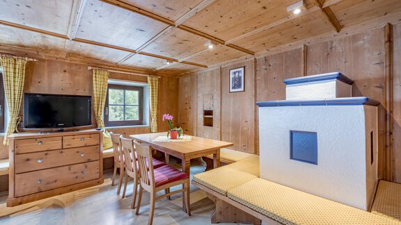 A cozy living space with wood paneling, a dining table, and chairs. The room features a television and a tiled stove. - Landhaus Peater's Hoamat - Sölden