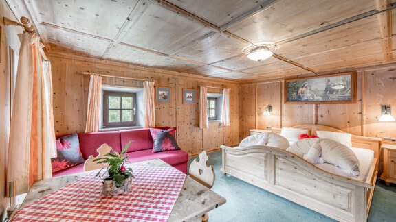 A cozy room with paneling, a sofa, and a bed. The table is covered with a red and white checked cloth, and a flower stands in the middle. - Landhaus Peater's Hoamat - Sölden
