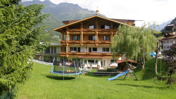 A cozy wooden house in a green landscape, surrounded by trees and mountains. The garden features a slide and a trampoline for children. - Pension Sportalm - Sölden