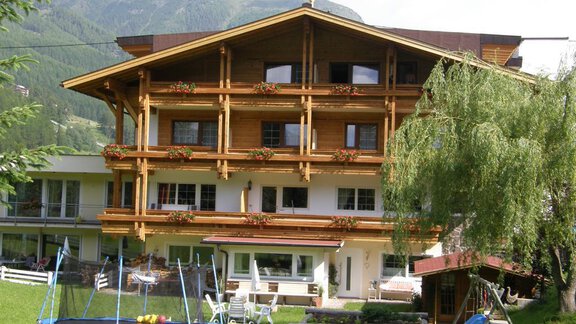 A cozy wooden house with multiple floors and balconies, surrounded by green grass and mountains. In the foreground, there is a trampoline and a swing. - Pension Sportalm - Sölden