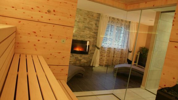 A modern wellness area with a wooden sauna and a cozy fireplace. Through large windows, the relaxation area is visible, offering a tranquil atmosphere. - Pension Sportalm - Sölden