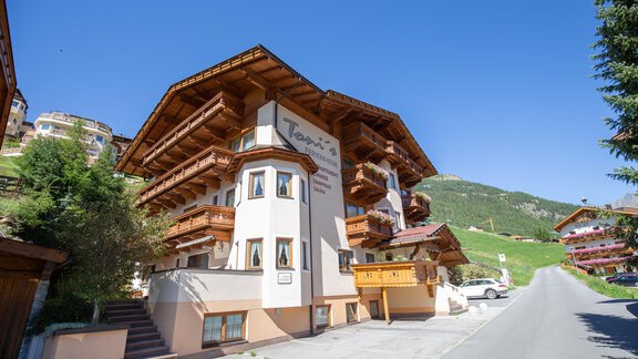 A modern building in alpine style with wooden cladding and balconies. The surroundings feature green hills and a clear blue sky. - Toni's Ferienheim - Sölden