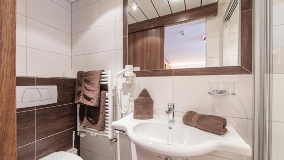 A modern bathroom with white tiles and wooden details. It is equipped with a toilet, a sink, and towels. - Vizensn Living - Sölden