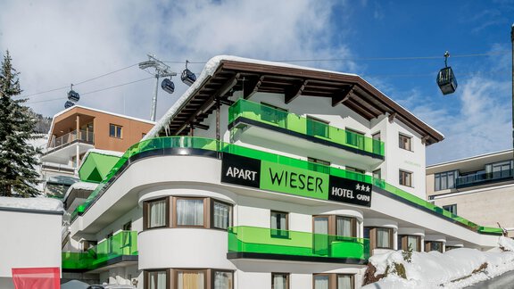 A modern hotel building with green accents is located in a snowy environment. In the background, ski lifts and clear skies are visible. - Apart Hotel Garni Wieser - Sölden