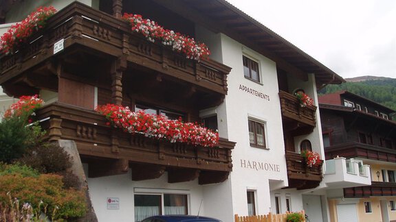 A charming building with a wooden balcony and colorful flowers. In addition, some cars and the surrounding nature are visible. - Appartement Harmonie - Sölden