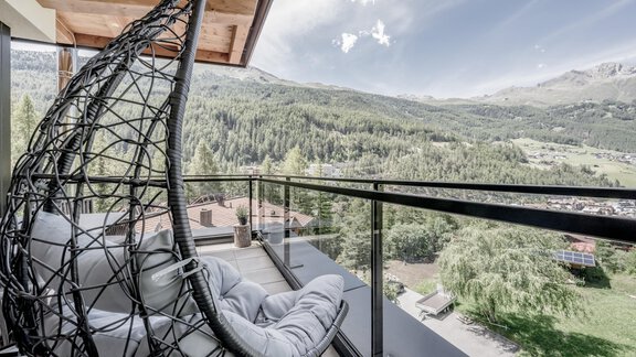 A modern rocking chair on a balcony overlooking a green mountain landscape. The sky is clear and the surroundings are calm and inviting. - CORSO living Aparthotel - Sölden