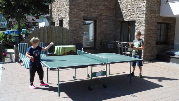 Two children are playing table tennis on a terrace. In the background, sunny trees and a building can be seen. - Ferienappartements Linserhof - Sölden | © Linserhof Ferienappartements
