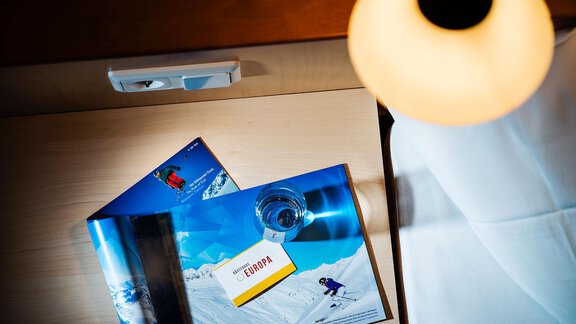 A table surface with tourist brochures on the topic of snow and skiing. Next to it, there is a glass of water and a lamp. - Gästehaus Europa - Sölden