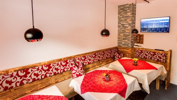 A cozy restaurant with red-clothed tables and modern pendant lights. The walls are designed with wood and stone, and a screen is mounted on the wall. - Haus Edmund - Sölden
