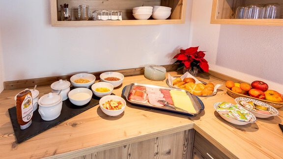 A lavish breakfast buffet with various bowls, cold cuts, cheese, and bread rolls on a wooden table. In the background, decorative elements and fresh fruit are visible. - Haus Edmund - Sölden