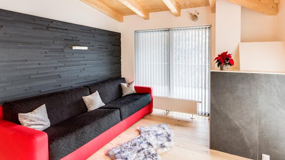 A modern living room with a red sofa and a black wall panel. Large windows provide plenty of daylight. - Haus Edmund - Sölden