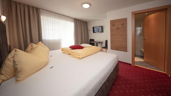 A cozy hotel room with a large double bed and soft pillows. The room is bright and inviting, with a desk and a bathroom in the background. - Haus Gondelblick - Sölden | © Falkner Georg