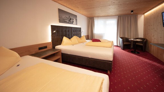 A cozy hotel room with two beds and yellow bedspreads. There is a table with chairs and large windows that let in plenty of light. - Haus Gondelblick - Sölden | © Falkner Georg