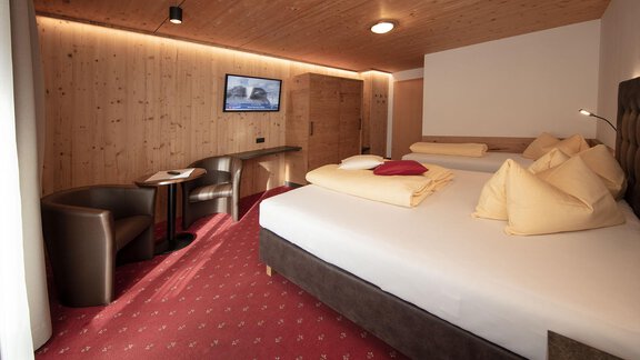 A modern hotel room with wooden paneling and red carpet. There are two beds, a television, and seating. - Haus Gondelblick - Sölden | © Falkner Georg