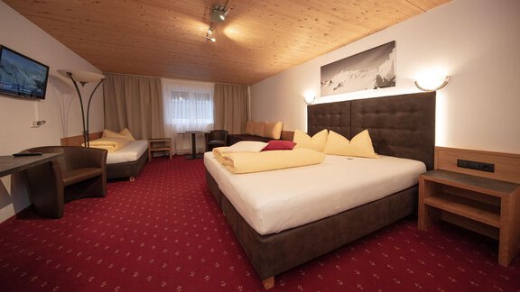 A modern hotel room with a large double bed, two single beds, and a cozy seating area. The decor is warm and inviting, with a wooden ceiling and soft lighting elements. - Haus Gondelblick - Sölden | © Falkner Georg