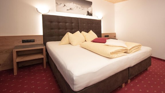 A cozy bedroom with a large bed and soft pillows. The walls are bright, and the floor is covered with a red carpet. - Haus Gondelblick - Sölden | © Falkner Georg