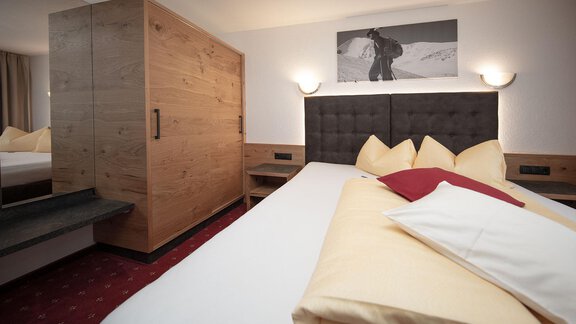 A modern hotel room with a large bed and elegant wooden paneling. In the background, there is a picture of a mountaineer hanging. - Haus Gondelblick - Sölden | © Falkner Georg
