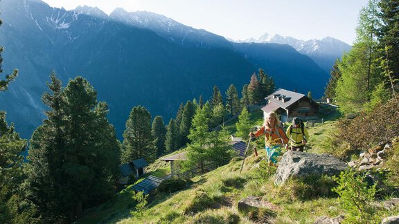 Two hikers are climbing up a mountain slope, surrounded by a beautiful forest landscape. In the background, majestic mountains and a mountain hut can be seen. - Haus Gondelblick - Sölden