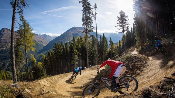 Two mountain bikers are riding on a winding trail through a forest. In the background, tall mountains and a clear sky are visible. - Haus Gondelblick - Sölden