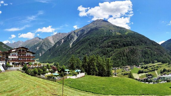 A picturesque mountain landscape with lush green and a bright blue sky. In the foreground, there is a hotel and in the background, majestic mountains rise up. - Pension Klaus - Sölden