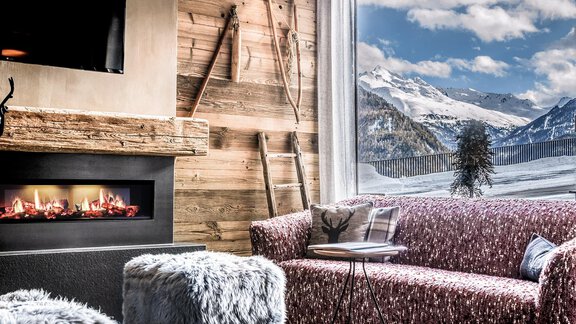 A cozy living room with a modern fireplace and a large window front. Outside, a snow-covered mountain landscape can be seen. - The Peak Sölden - Sölden