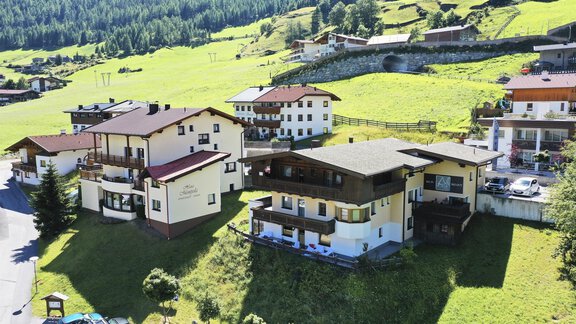 A tranquil landscape with several houses on a green meadow. In the background, gentle hills and trees can be seen. - A CASA Appartements Juwel - Sölden
