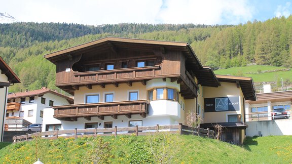 A typical alpine house with wooden balconies and a large window front. It is located surrounded by green meadows and mountains. - A CASA Appartements Juwel - Sölden