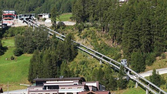 A mountain railway travels along a green hilly landscape. In the foreground, modern buildings and dense forests are visible. - A CASA Appartements Juwel - Sölden