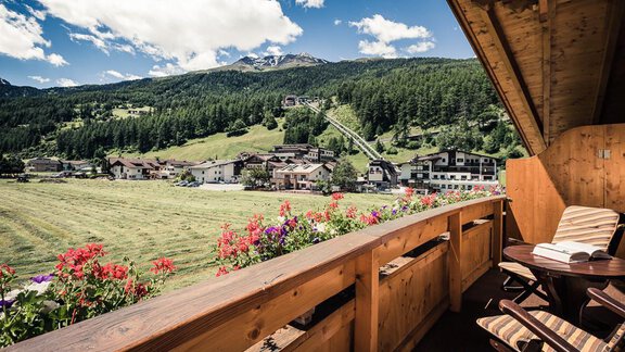 A view from a balcony overlooking a picturesque landscape with mountains and a small village. Colorful flowers adorn the railing, and the sky is covered with few clouds. - Andre Arnold - Boutique Hotel - Sölden | © Rudi Wyhlidal