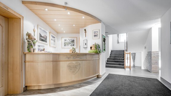 A bright reception hall with a wood reception desk and modern pictures on the wall. In the background, stairs can be seen leading to other areas. - Appartementhaus Kathrin - Sölden