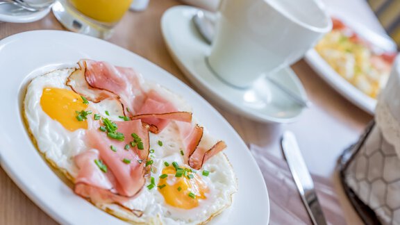 A plate with fried eggs topped with ham and fresh chives. In the background, a drink and a cup are visible. - Appartementhaus Kathrin - Sölden