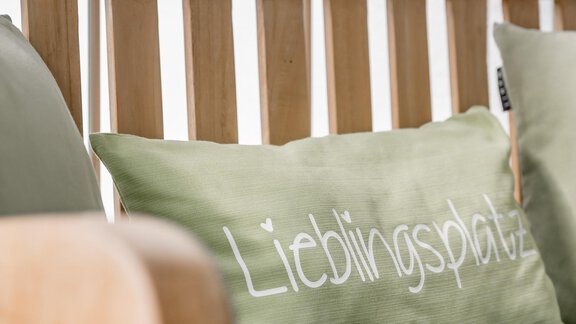A wooden bench with a gray cushion and a green cushion that reads "Favorite Spot". The bench is in a bright, inviting room. - Appartementhaus Kathrin - Sölden