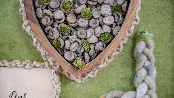 A heart-shaped wooden bowl contains small stones and green plants. Next to it is a braided ribbon made from various materials. - Appartementhaus Kathrin - Sölden