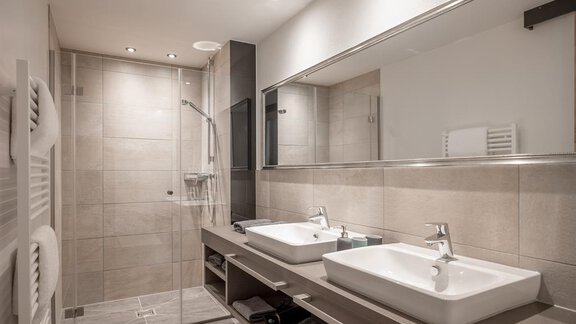 A modern bathroom with two sinks and a clear mirror. The shower is separated by a glass wall and the walls are in neutral tones. - CORSO living Aparthotel - Sölden