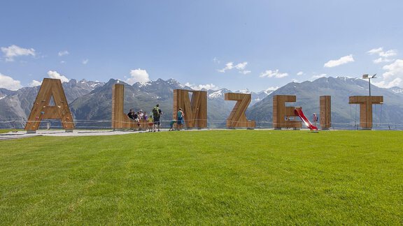 A large wooden sculpture with the inscription "ALMZEIT" in front of an impressive mountain landscape. The grass is green, and the sky is blue with a few clouds. - CORSO living Aparthotel - Sölden
