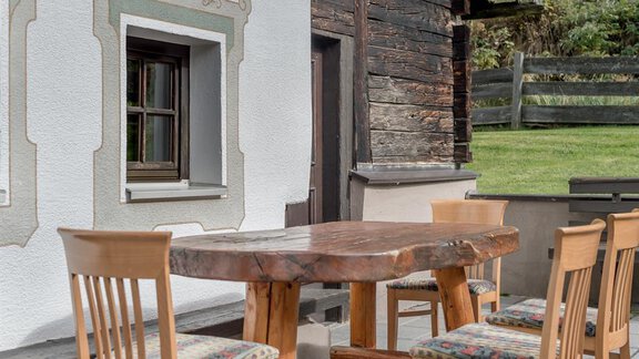 A rustic wooden table with chairs stands in front of a traditional building. In the background, green meadows and a wooden fence can be seen. - Landhaus Peater's Hoamat - Sölden